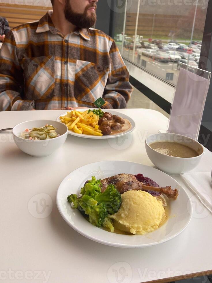 Millennial man in a plaid shirt sits at a white table with a hearty meal in front of him, featuring mashed potatoes, broccoli and big portion of protein. photo