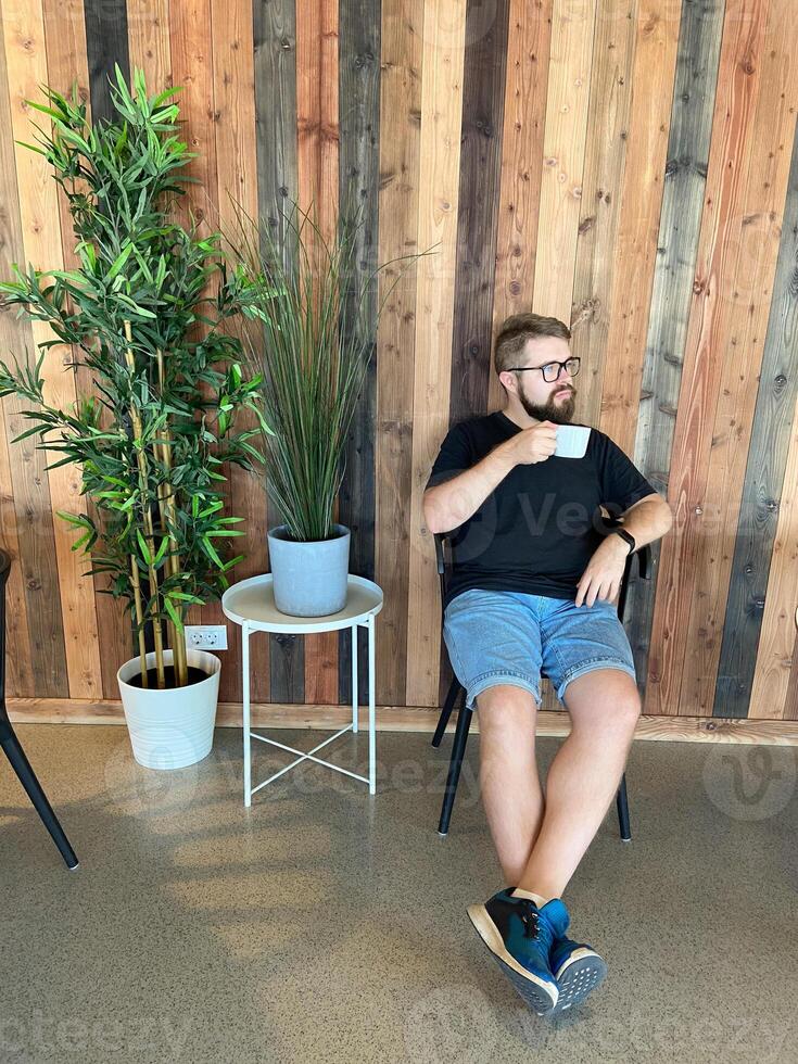 Bearded man in casual attire sits on a chair, holding a cup of coffee, against a stylish wooden wall with potted plants. Relaxed and modern atmosphere, perfect for enjoying a peaceful moment photo