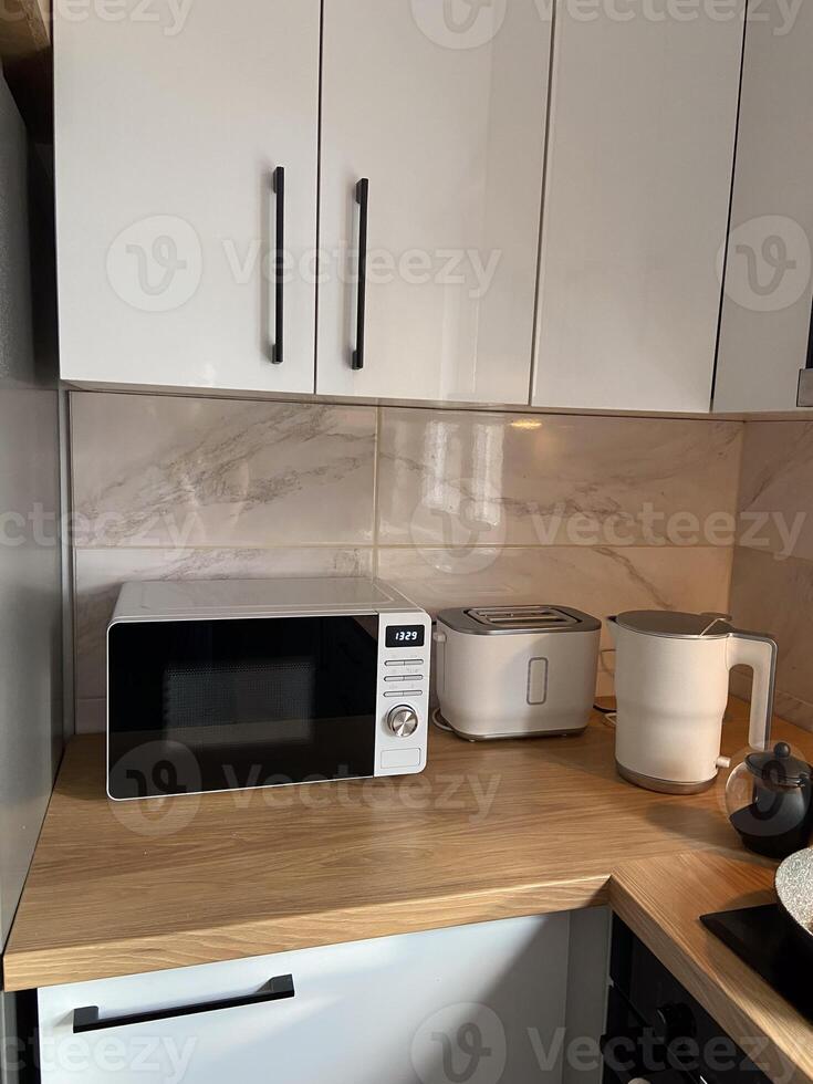 Modern kitchen corner features a microwave, a toaster, and an electric kettle on a wood countertop. Minimalist design with sleek white cabinets photo
