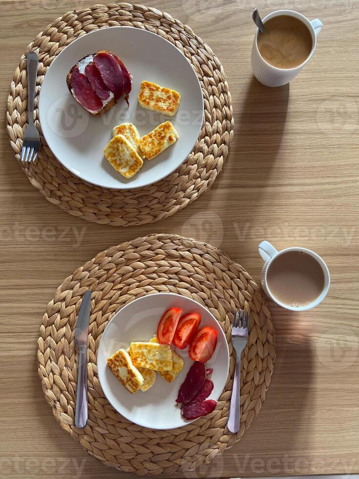 Cozy breakfast setting features plates with cheese, tomatoes, and cured meat, complemented by cups of coffee. Woven placemats and wooden table provide a warm, inviting ambiance photo