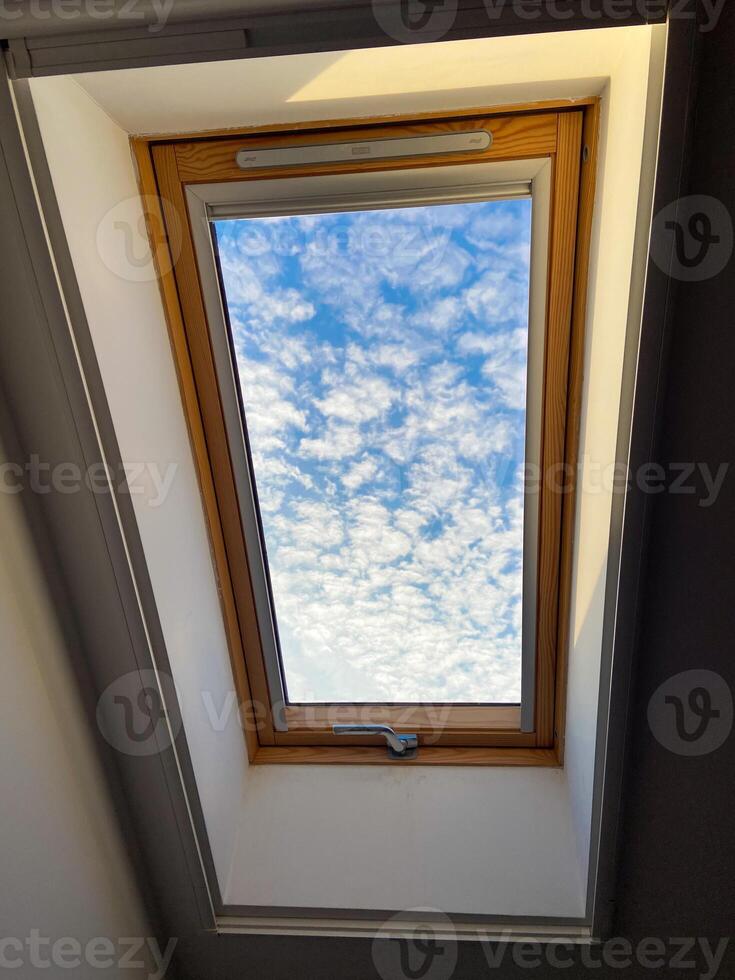 Clear sky view through a skylight reveals a vibrant array of clouds, offering a serene glimpse of outdoors. Wooden frame adds warmth and contrast to the bright blue sky photo