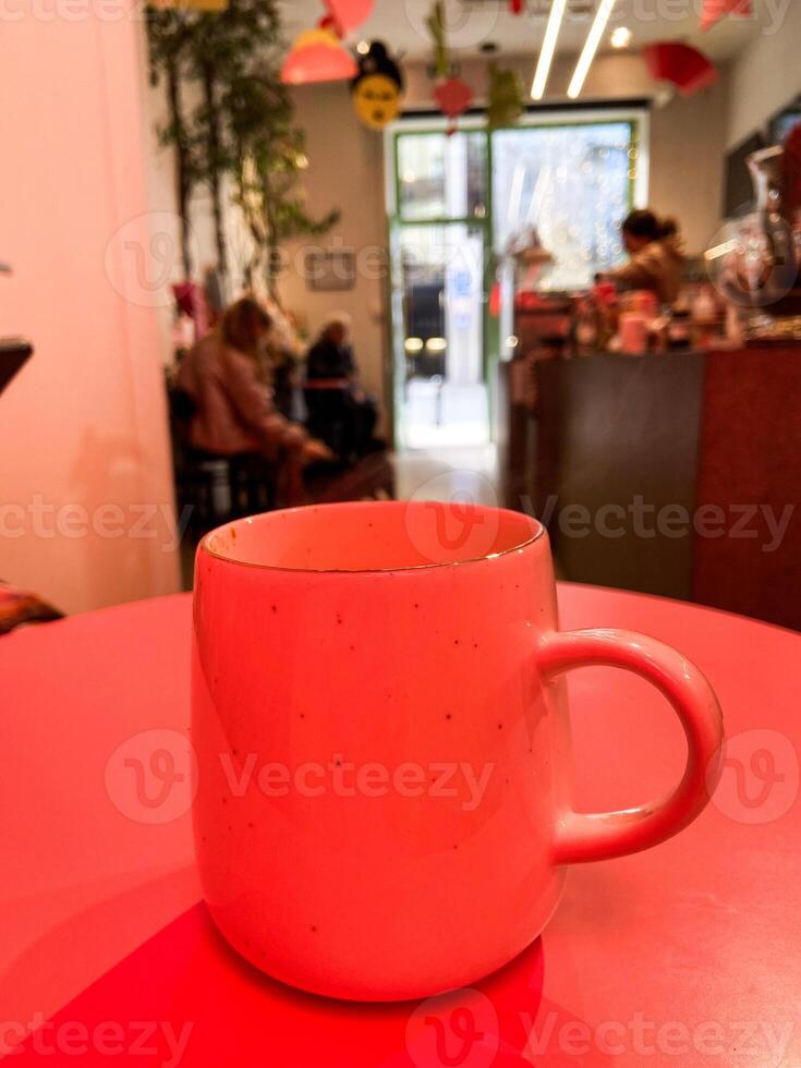 Mug with hot winter drink on a table in cozy cafe with bright red light. Blurred figures, plants, and festive Christmas decorations create warm and inviting atmosphere. Coffee shop photo