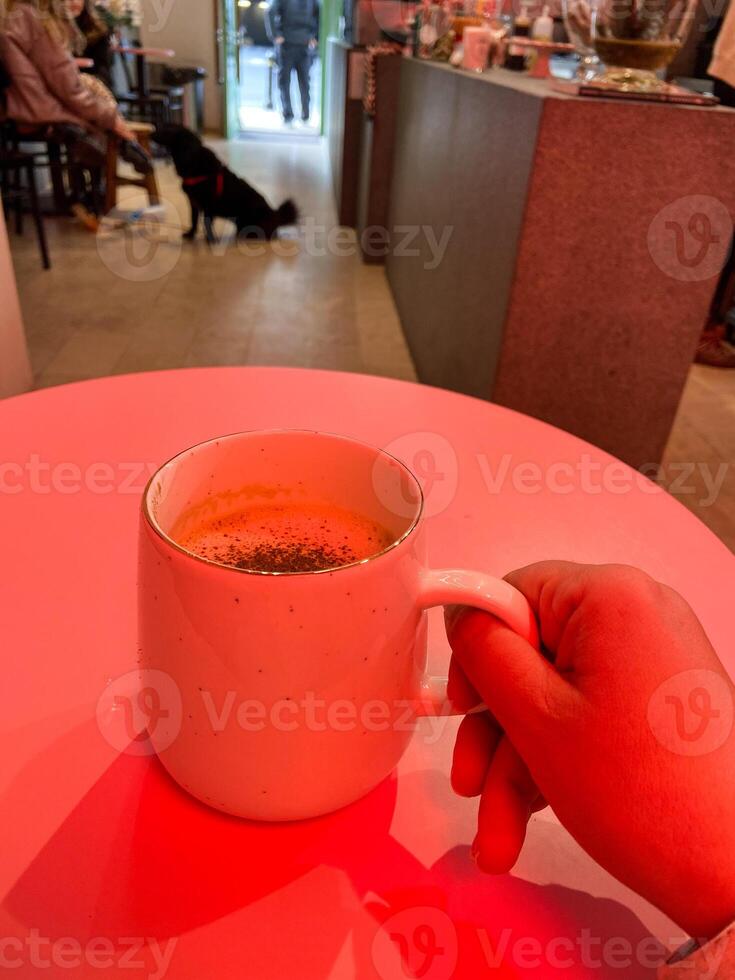 Mug with hot winter drink on a table in cozy cafe with bright red light. Blurred figures, plants, and festive Christmas decorations create warm and inviting atmosphere. Coffee shop photo