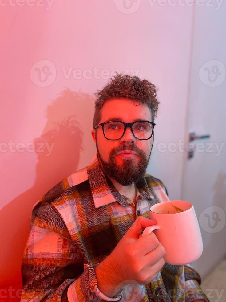 Millennial man with glasses drinks from a large mug coffee. Warm red light in coffee shop creating a cozy and relaxed atmosphere. Winter cold Christmas season photo