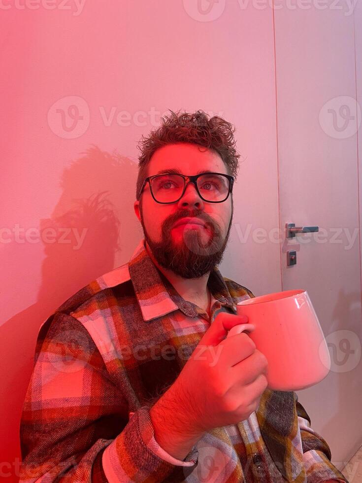 Millennial man with glasses drinks from a large mug coffee. Warm red light in coffee shop creating a cozy and relaxed atmosphere. Winter cold Christmas season photo