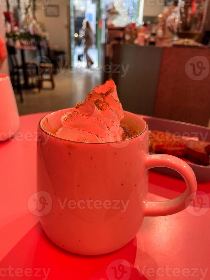 Mug with hot winter drink on a table in cozy cafe with bright red light. Blurred figures, plants, and festive Christmas decorations create warm and inviting atmosphere. Coffee shop photo