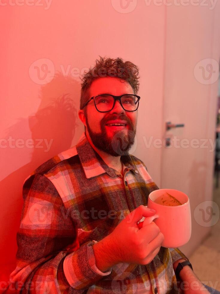 Millennial man with glasses drinks from a large mug coffee. Warm red light in coffee shop creating a cozy and relaxed atmosphere. Winter cold Christmas season photo