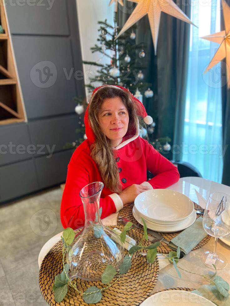 Millennial woman dressed in a Santa outfit sits at a beautifully set holiday table, surrounded by festive decorations and a Christmas tree. Cozy scene illuminated photo
