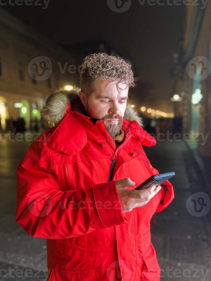 Millennial man in vivid red parka stands on city street at night, engrossed in their smartphone. Illuminated by the warm glow of city lights, capturing essence of urban life and digital connectivity. photo