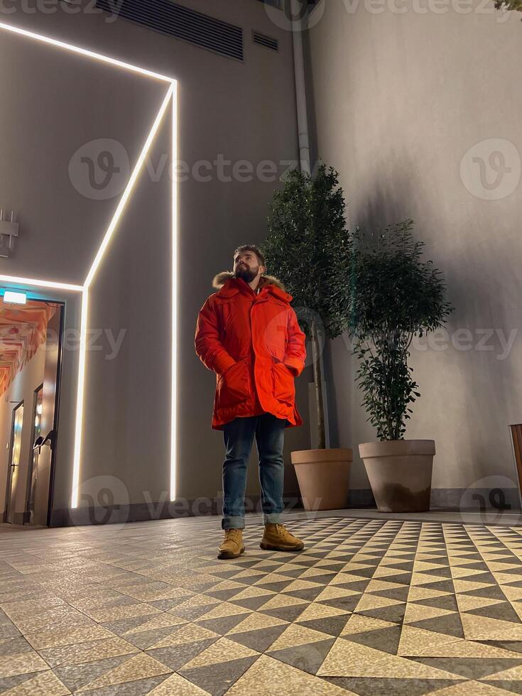 Millennial man stands confidently in a modern space, wearing a vibrant orange parka, surrounded by geometric patterns and ambient lighting. Blend of contemporary style and cozy warmth photo