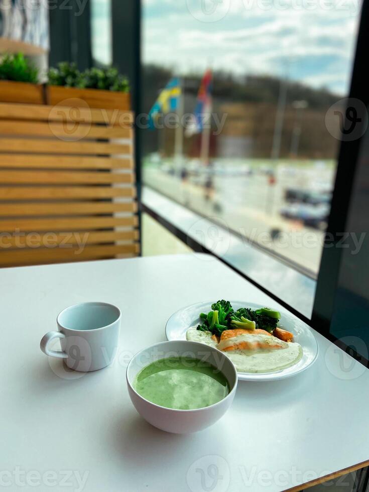 Healthy meal featuring a bowl of green soup and a plate with broccoli and fish is set on a white table by a large window. Peaceful dining atmosphere photo