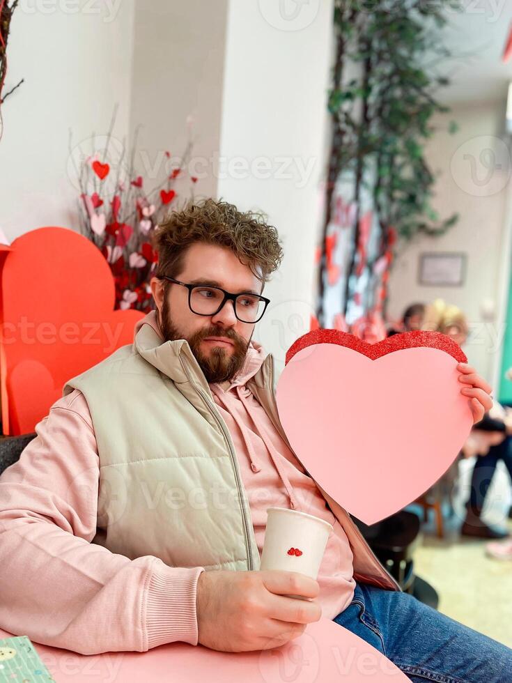 Smiling man holds a large pink heart on Valentine day while sitting at festively decorated table, capturing spirit of love and warmth in a cozy setting. Cheerful festive atmosphere evoke photo