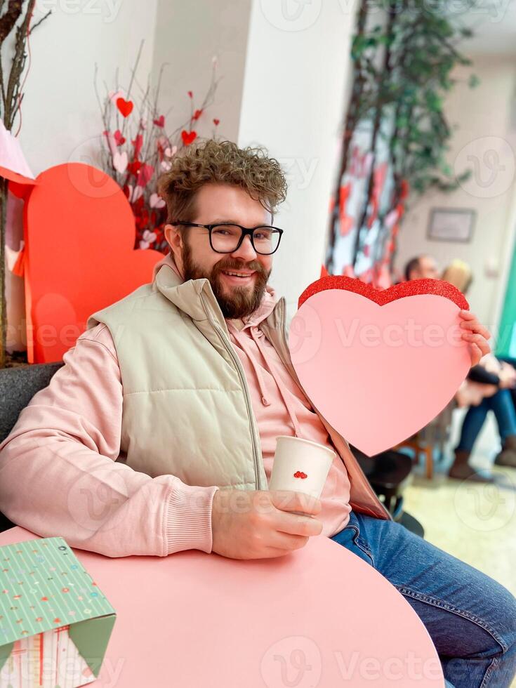 Smiling man holds a large pink heart on Valentine day while sitting at festively decorated table, capturing spirit of love and warmth in a cozy setting. Cheerful festive atmosphere photo