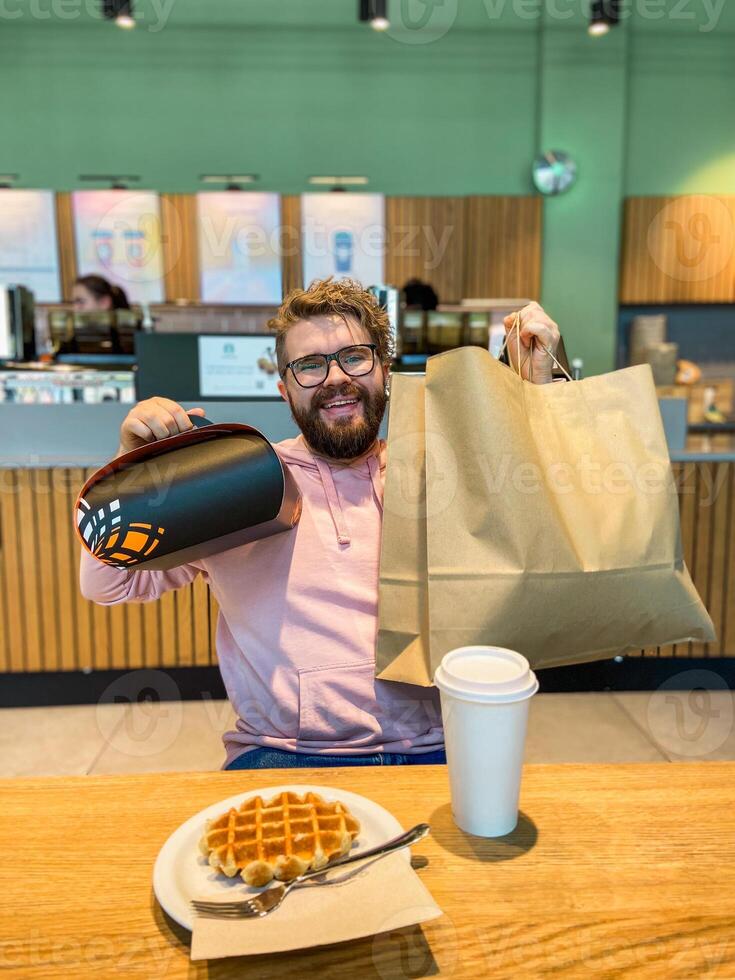 Surprised man sits at a cafe table with a waffle, holding a large shopping bag and a takeout gift box. Moment of unexpected delight and enjoyment in a modern coffee shop photo