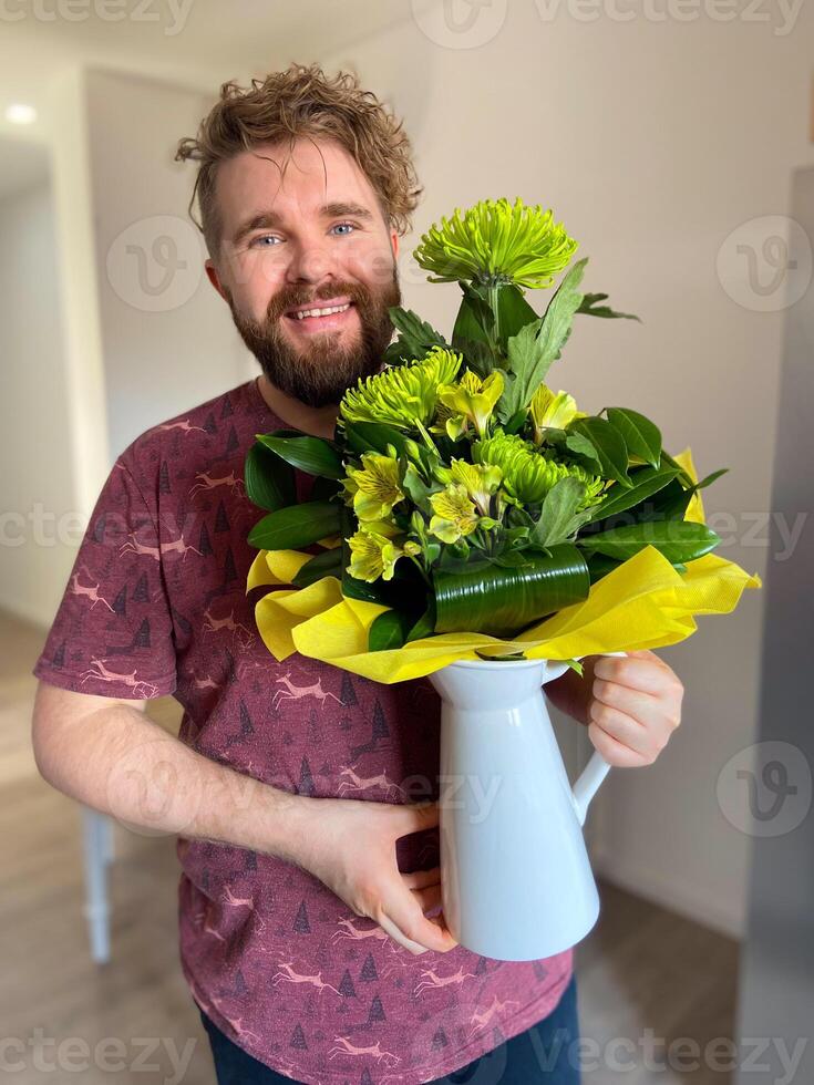 Bearded smiling man holds a vibrant bouquet of green and yellow flowers in a white pitcher, radiating warmth and happiness. Holidays and birthday concept photo