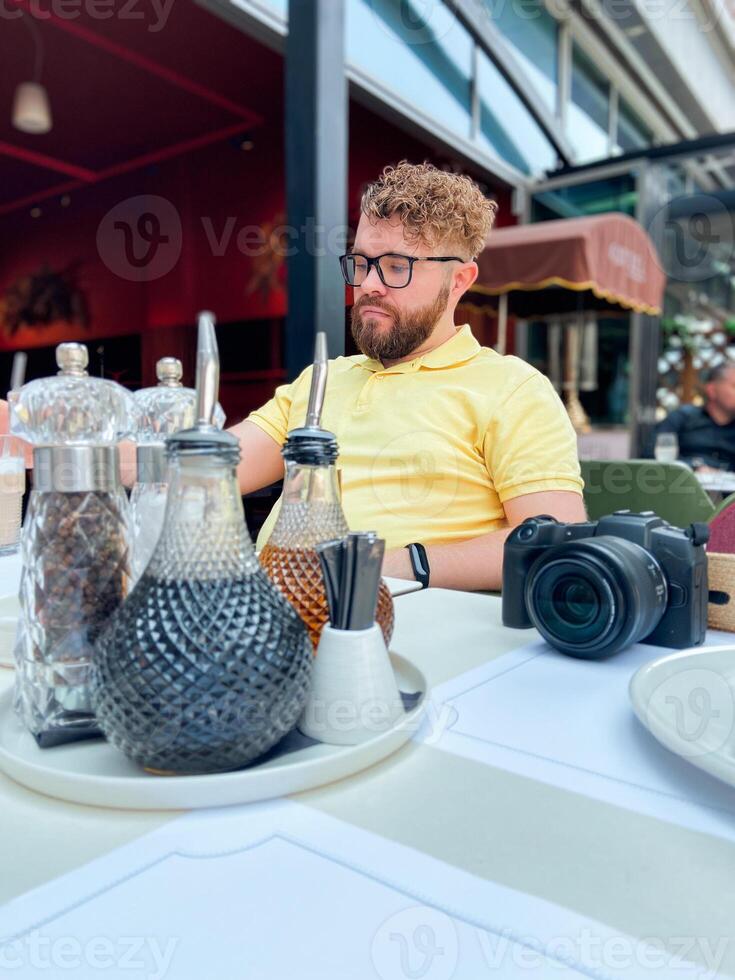 Millennial man photographer in a yellow shirt sits at an outdoor cafe, camera standing in front of him. Relaxed and enjoying a quiet moment at the restaurant, possibly working or reading. photo