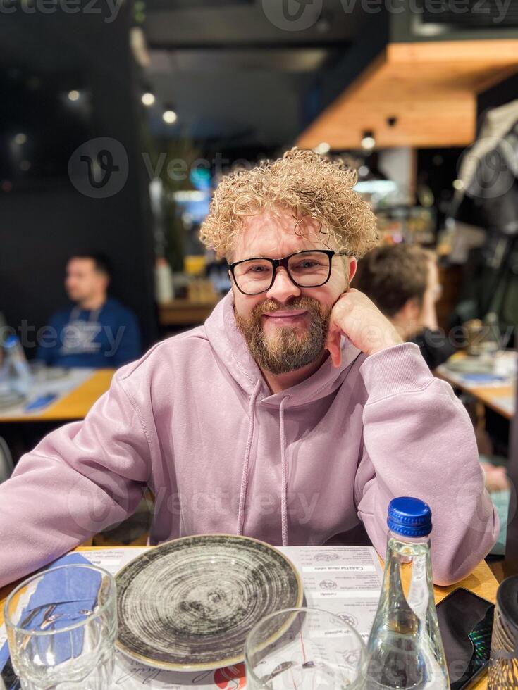 Curly smiling man in a casual hoodie sits at a restaurant table, radiating warmth and friendliness. glasses and a water bottle are neatly arranged, suggesting a cozy dining atmosphere photo