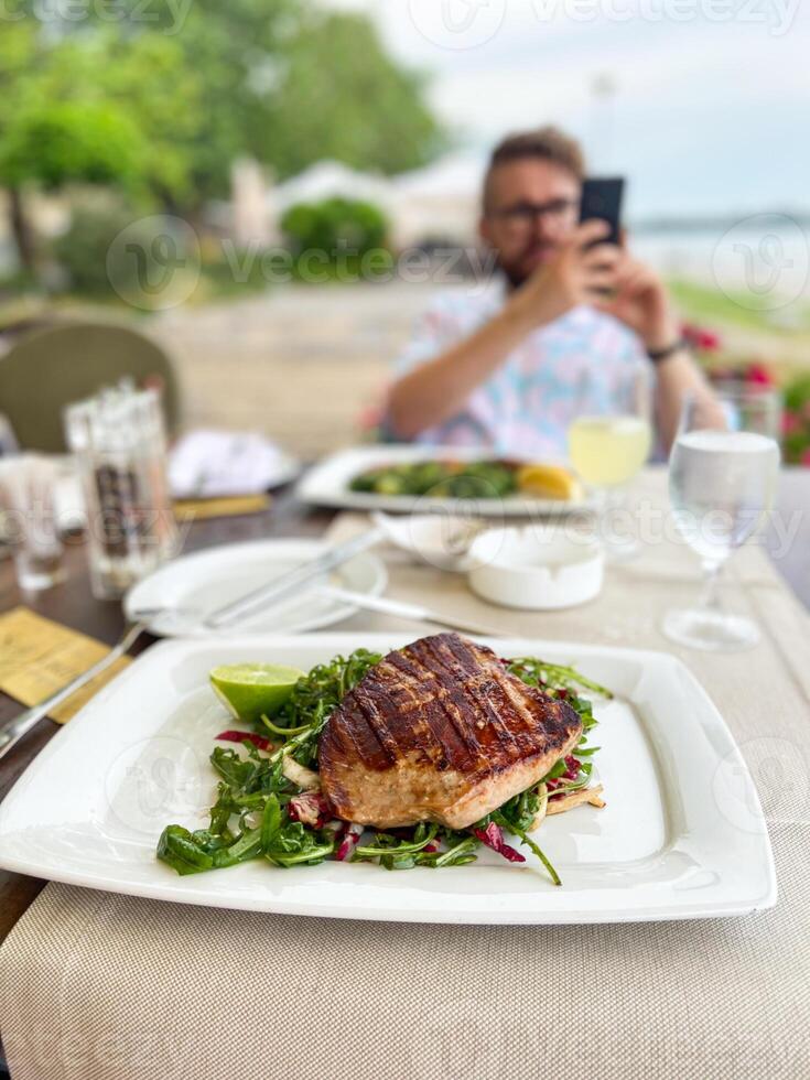 Close-up grilled fish on plate with fresh salad while man in background uses smartphone at outdoor restaurant. Casual dining experience by the waterfront photo