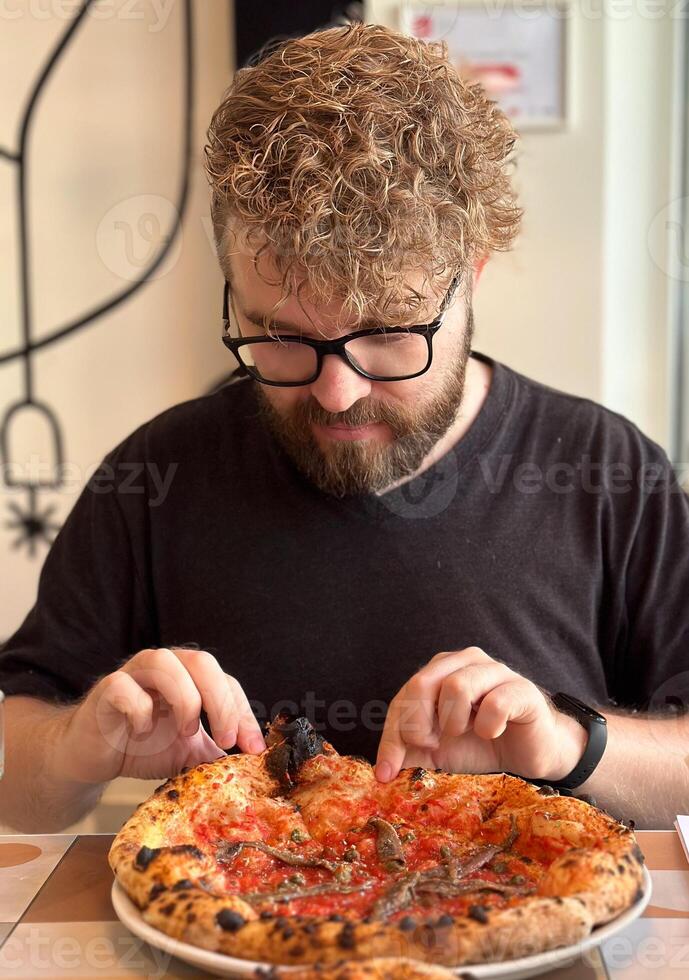 Millennial man with curly hair and glasses looking on a delicious-looking pizza in restaurant. Moment of culinary anticipation and simple joys of food photo