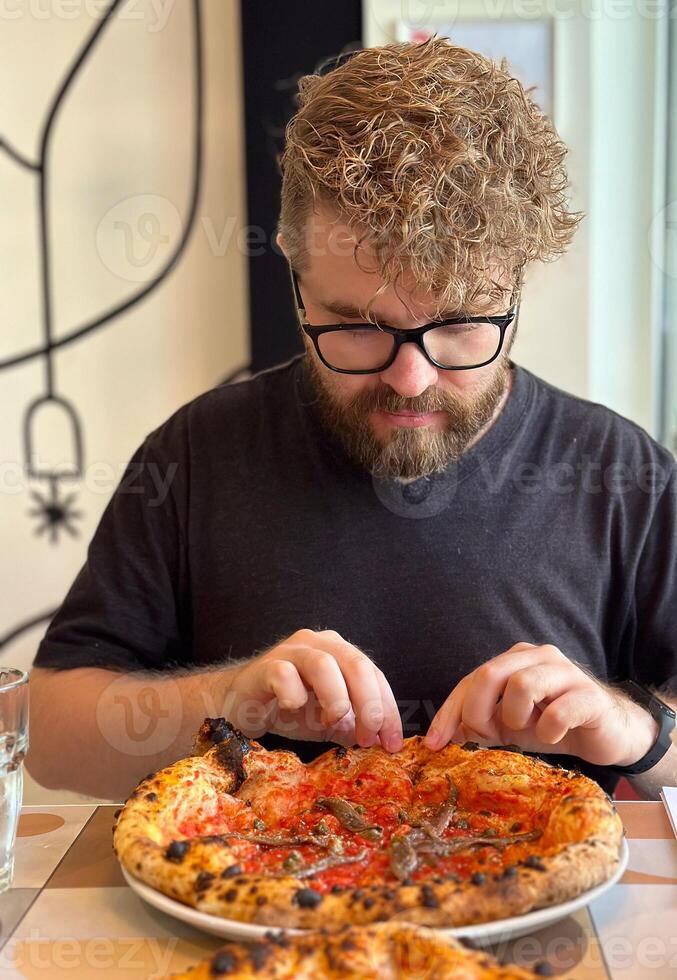 Millennial man with curly hair and glasses looking on a delicious-looking pizza in restaurant. Moment of culinary anticipation and simple joys of food photo
