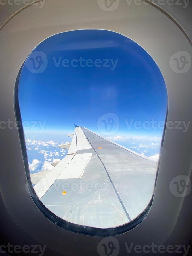 View of airplane wing through window at high altitude, showcasing clear blue sky and clouds far below. Serene beauty of flight and the expansive view of the horizon from above. photo