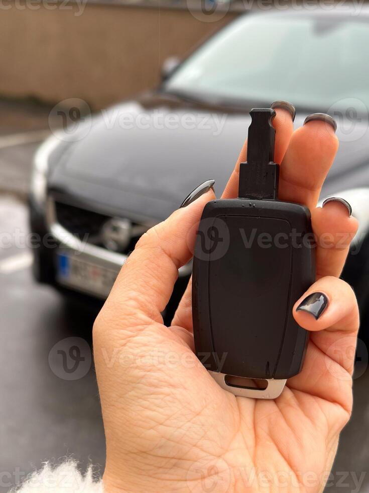 Woman's hand holding a car key fob in front of a black modern car on a rainy day. Fluffy jacket highlights the close-up shot of the key, ready to unlock the vehicle. photo