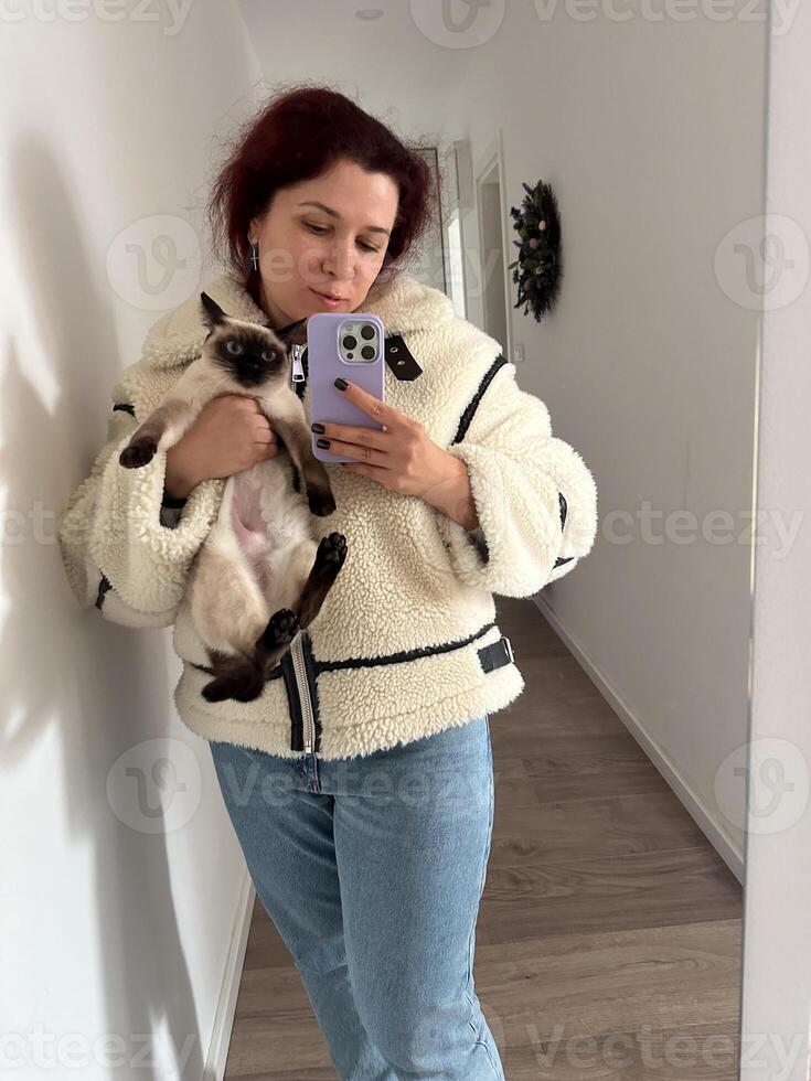 Cozy moment captured as woman holds a Siamese cat and takes a mirror selfie. Dressed in a fluffy jacket and holding smartphone, warm and intimate scene is framed by the hallway background photo