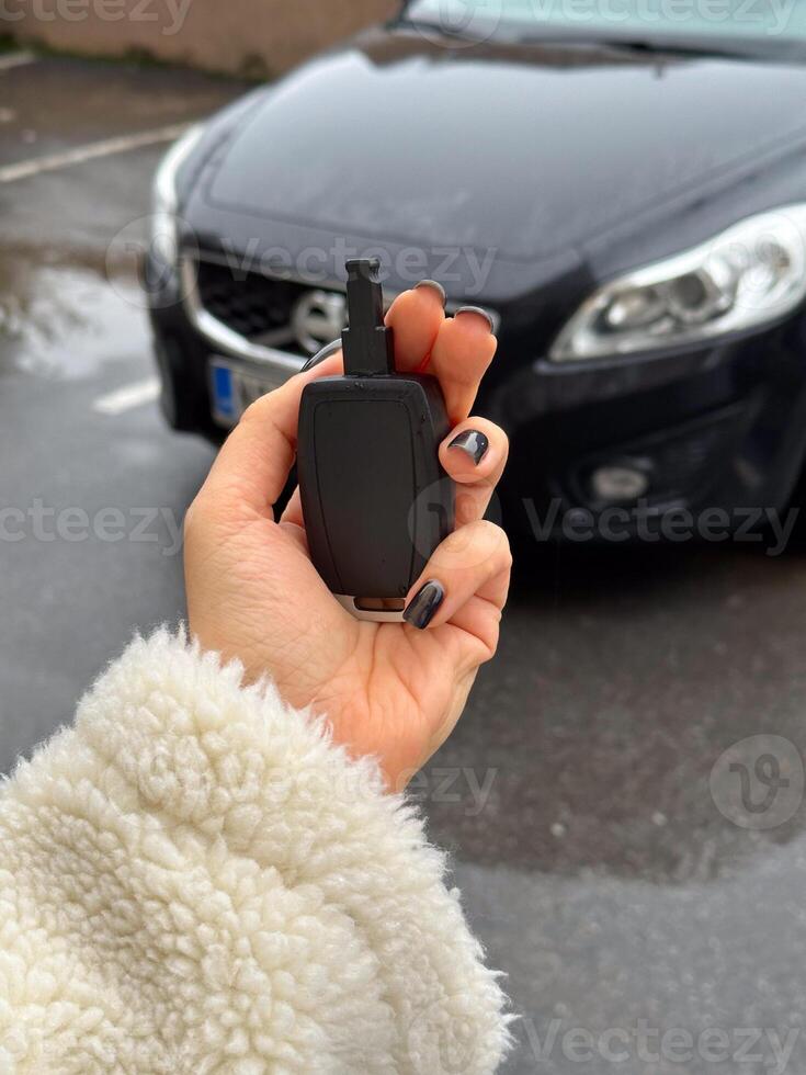 Woman's hand holding a car key fob in front of a black modern car on a rainy day. Fluffy jacket highlights the close-up shot of the key, ready to unlock the vehicle. photo