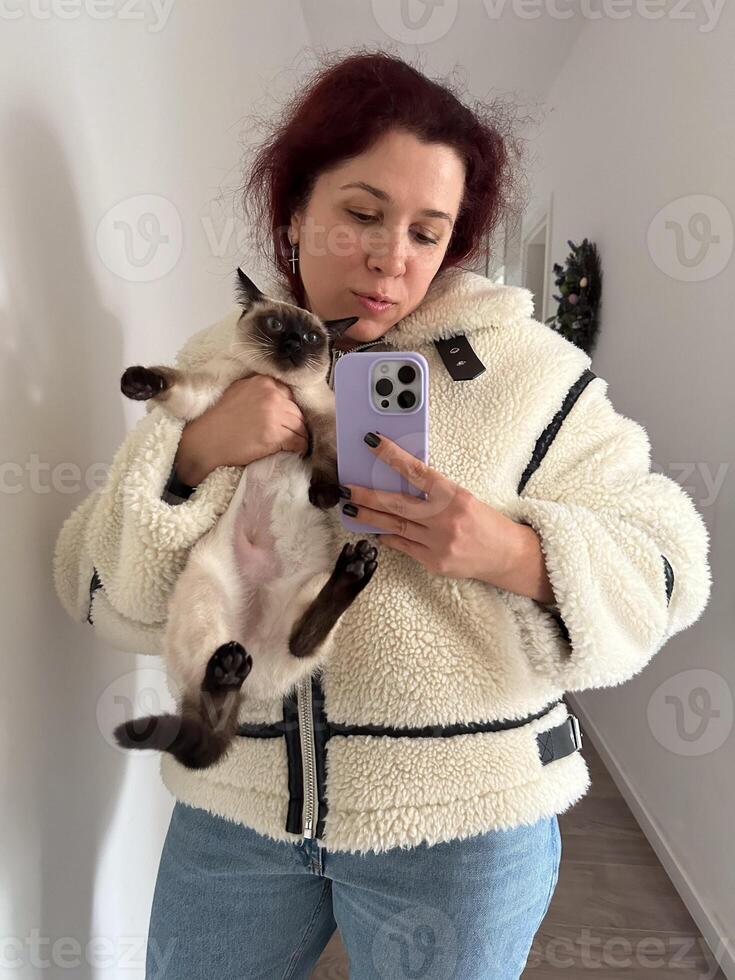 Cozy moment captured as woman holds a Siamese cat and takes a mirror selfie. Dressed in a fluffy jacket and holding smartphone, warm and intimate scene is framed by the hallway background photo