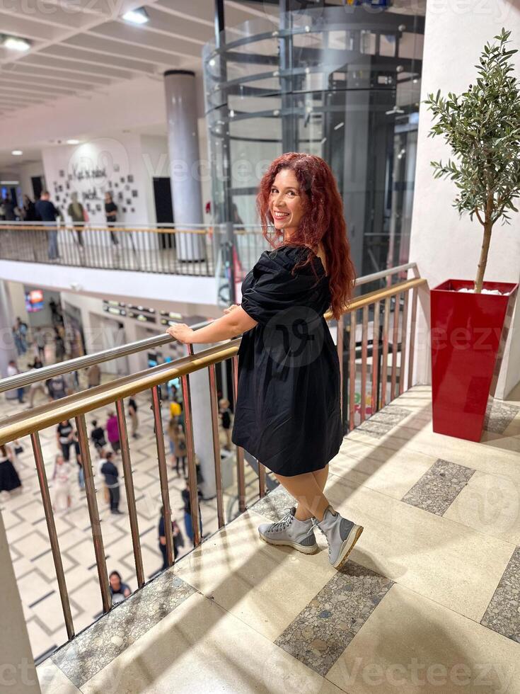 Woman posing indoors in a stylish black dress, dressed fashionably and confidently next to a railing and a red planter. Modern fashion and elegance in an urban setting photo