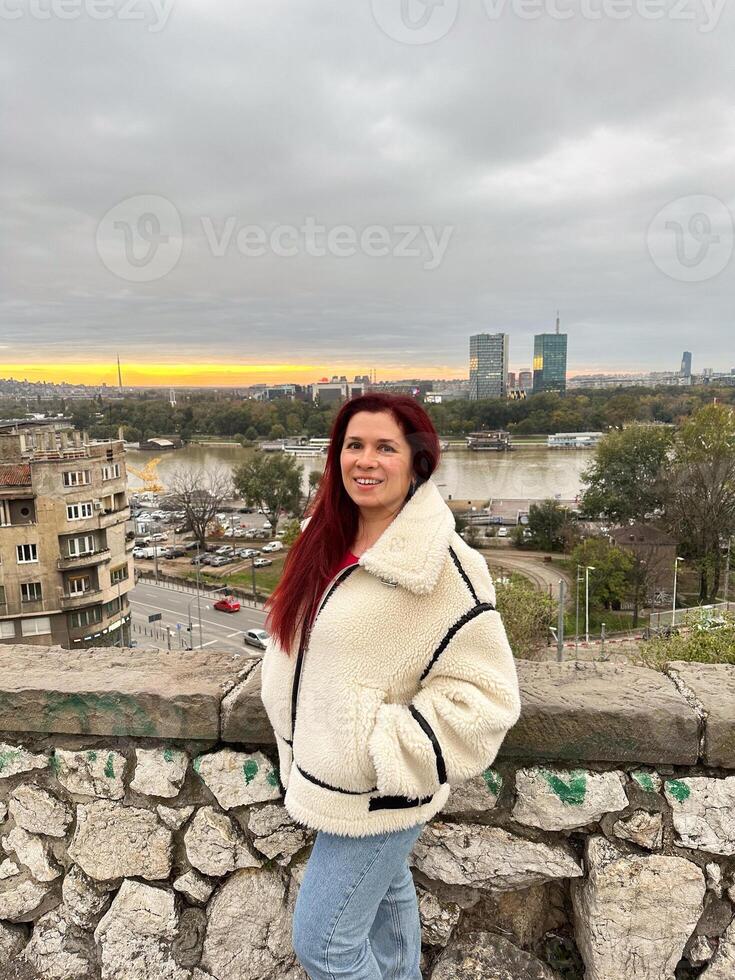 Woman in a cozy jacket overlooks a serene river and urban skyline at sunset in autumn season. Scene captures a moment of tranquility and reflection amidst an expansive cityscape photo