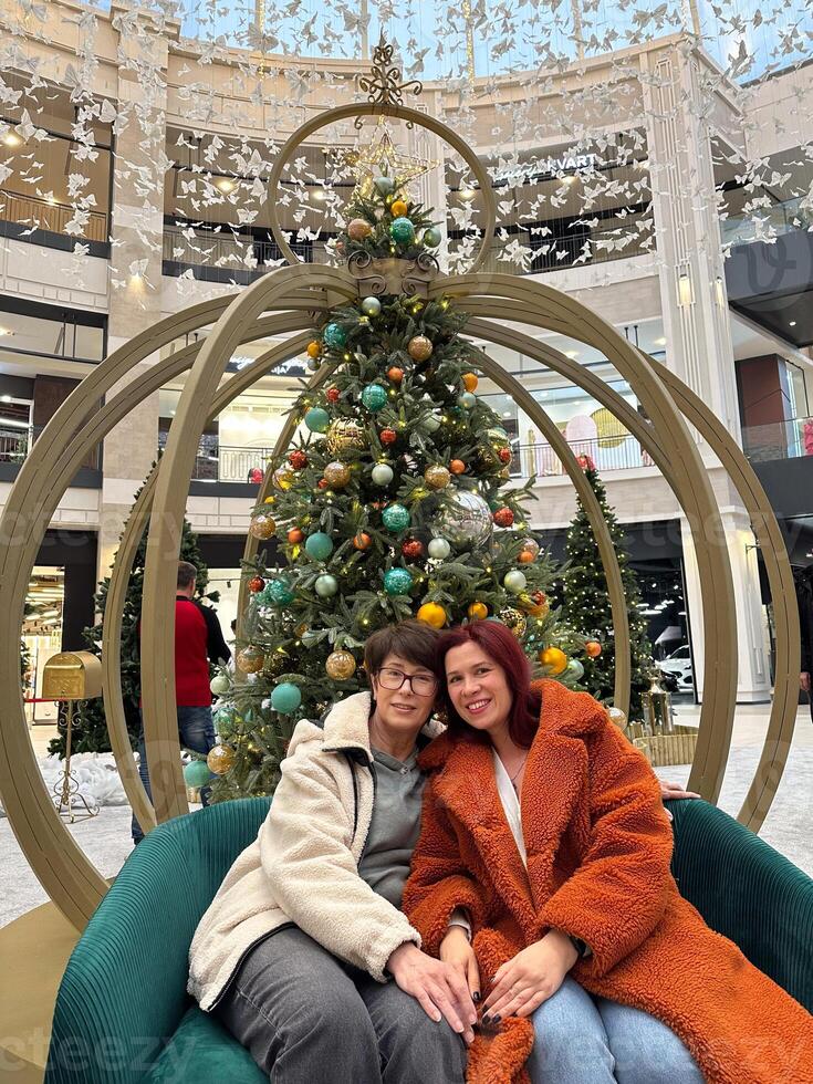 Mother and daughter sit comfortably on a stylish bench in front of decorated Christmas tree in a mall. the festive setting and their warm clothing create a cozy and joyful holiday atmosphere photo