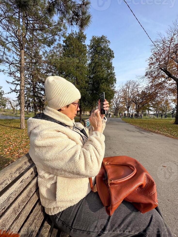 Woman taking a photo in a park on a sunny autumn day, moments of leisure and connection with nature amidst fall cold scenery