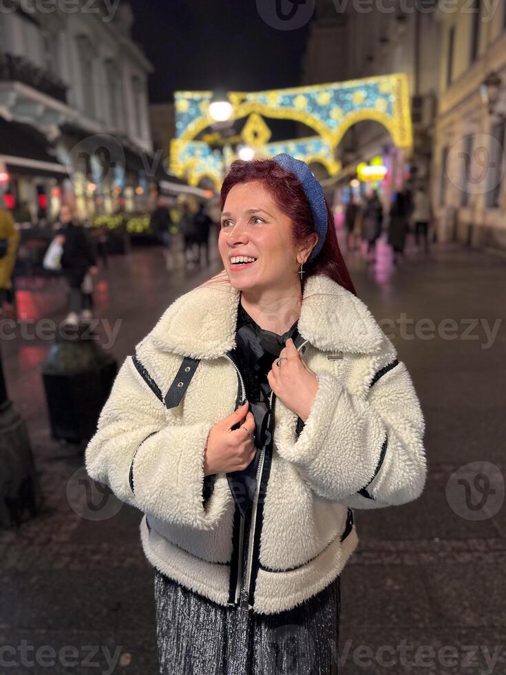 Millennial woman standing outdoors at Christmas night, wrapped in a cozy shearling jacket. the city lights in the background create a warm and Christmas holidays atmosphere, enhancing the peaceful photo