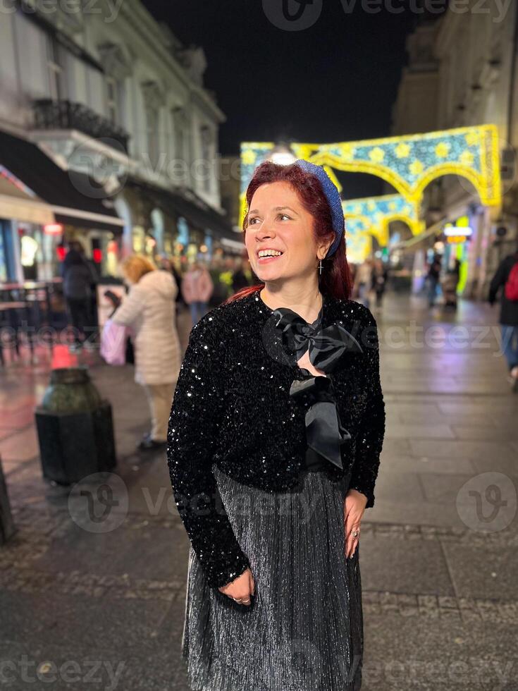 Millennial woman standing outdoors at Christmas night,. City lights in the background create a warm and Christmas holidays atmosphere, enhancing the peaceful and confident expression on her face photo
