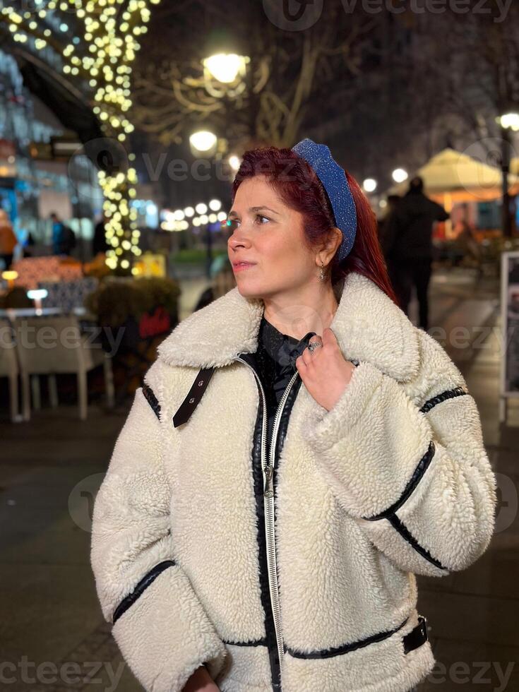 Millennial woman standing outdoors at Christmas night, wrapped in a cozy shearling jacket. the city lights in the background create a warm and Christmas holidays atmosphere, enhancing photo