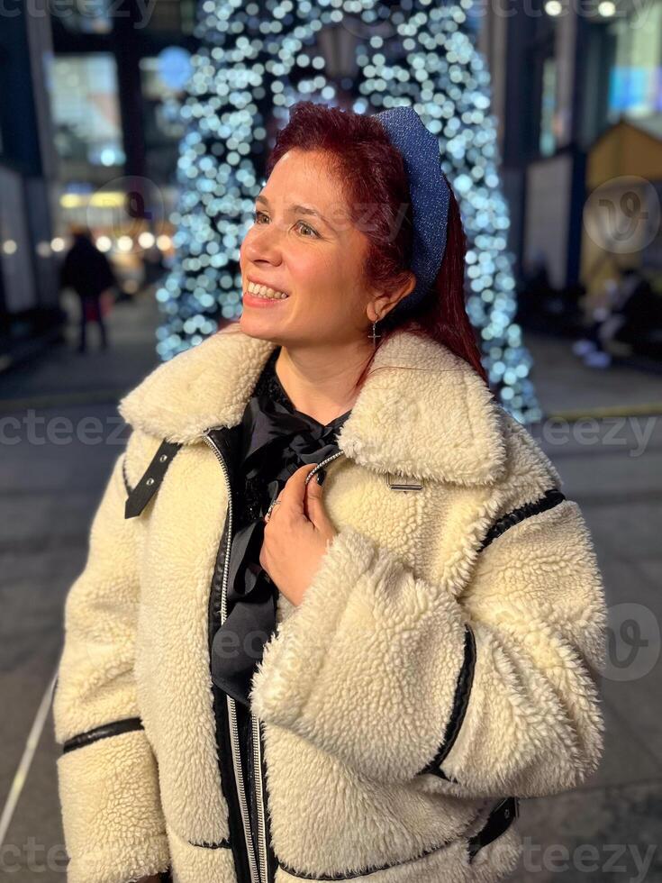 Millennial woman standing outdoors at Christmas night, wrapped in a cozy shearling jacket. the city lights in the background create a warm and Christmas holidays atmosphere, photo