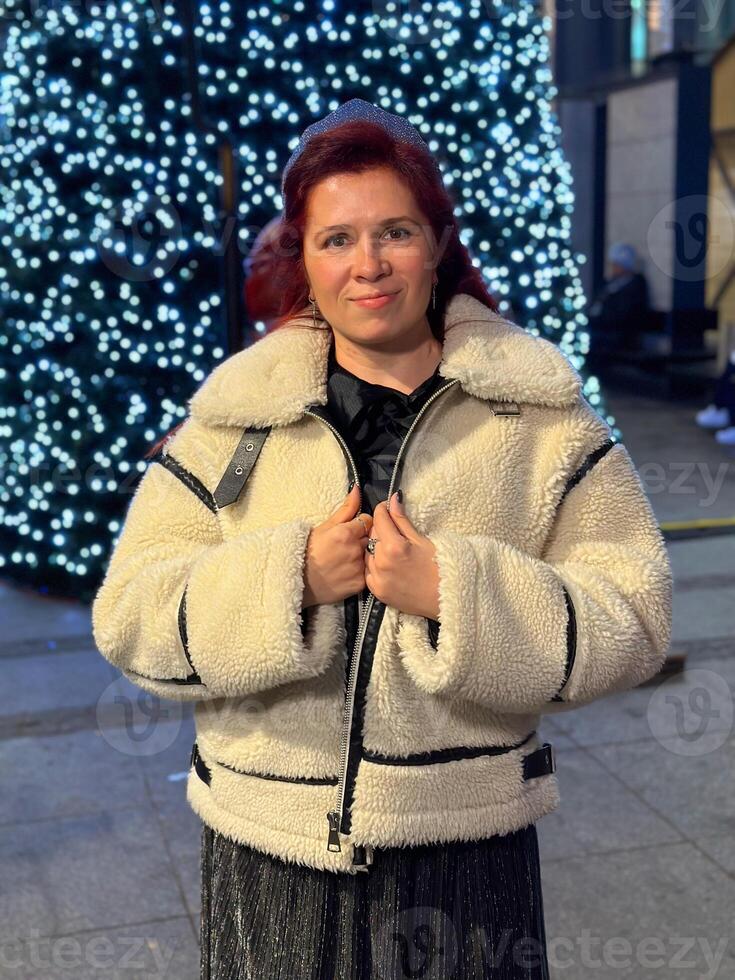 Millennial woman standing outdoors at Christmas night, wrapped in a cozy shearling jacket. the city lights in the background create a warm and Christmas holidays atmosphere, photo