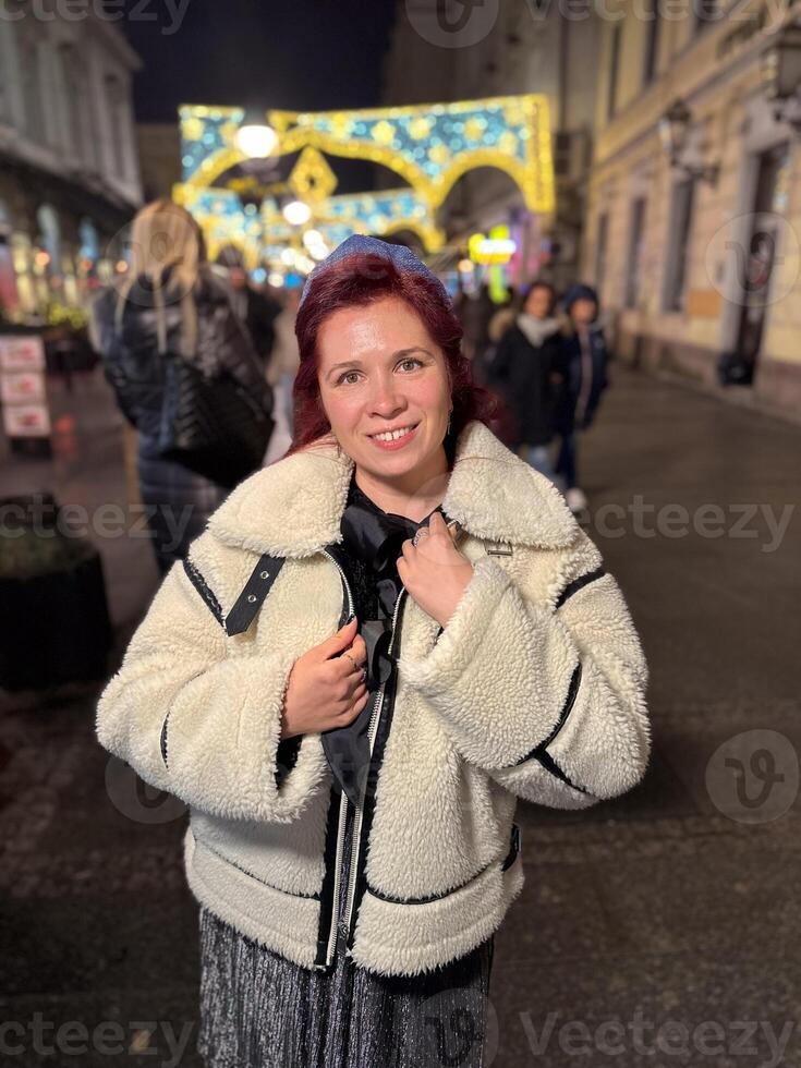 Millennial woman standing outdoors at Christmas night, wrapped in a cozy shearling jacket. the city lights in the background create a warm and Christmas holidays atmosphere, enhancing the peaceful photo
