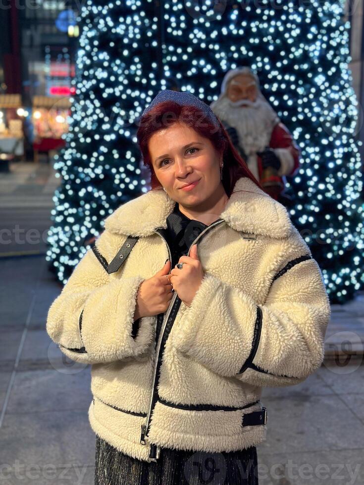 Millennial woman standing outdoors at Christmas night, wrapped in a cozy shearling jacket. the city lights in the background create a warm and Christmas holidays atmosphere, photo