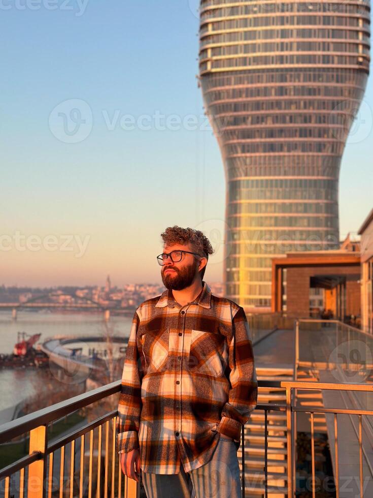 Man stands thoughtfully on a balcony, overlooking a river with a modern skyscraper in Belgrade city during a serene sunset. the warm glow of the evening light casts photo