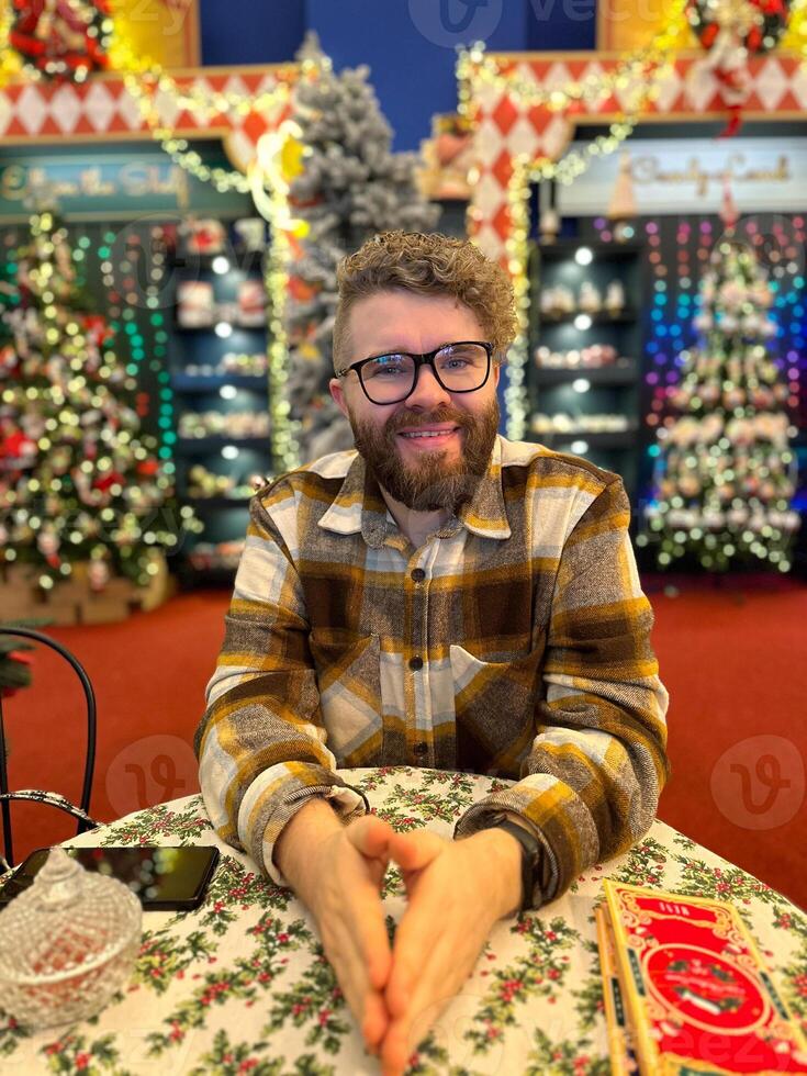 Millennial man sits cheerfully at a festively decorated table, surrounded by Christmas decorations in a cozy indoor setting. Vibrant colors and cheerful atmosphere reflect photo