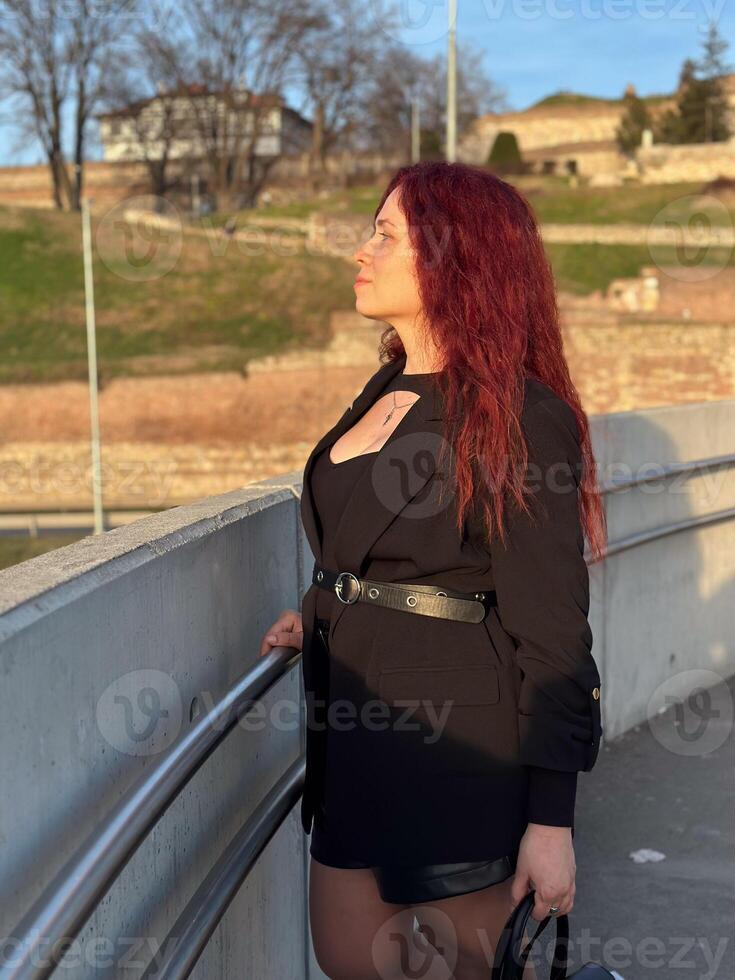 Millennial woman stands confidently in sunlight, with vibrant red hair flowing and dressed in a chic black outfit. Background of a clear blue sky and distant scenery enhances the dynamic photo