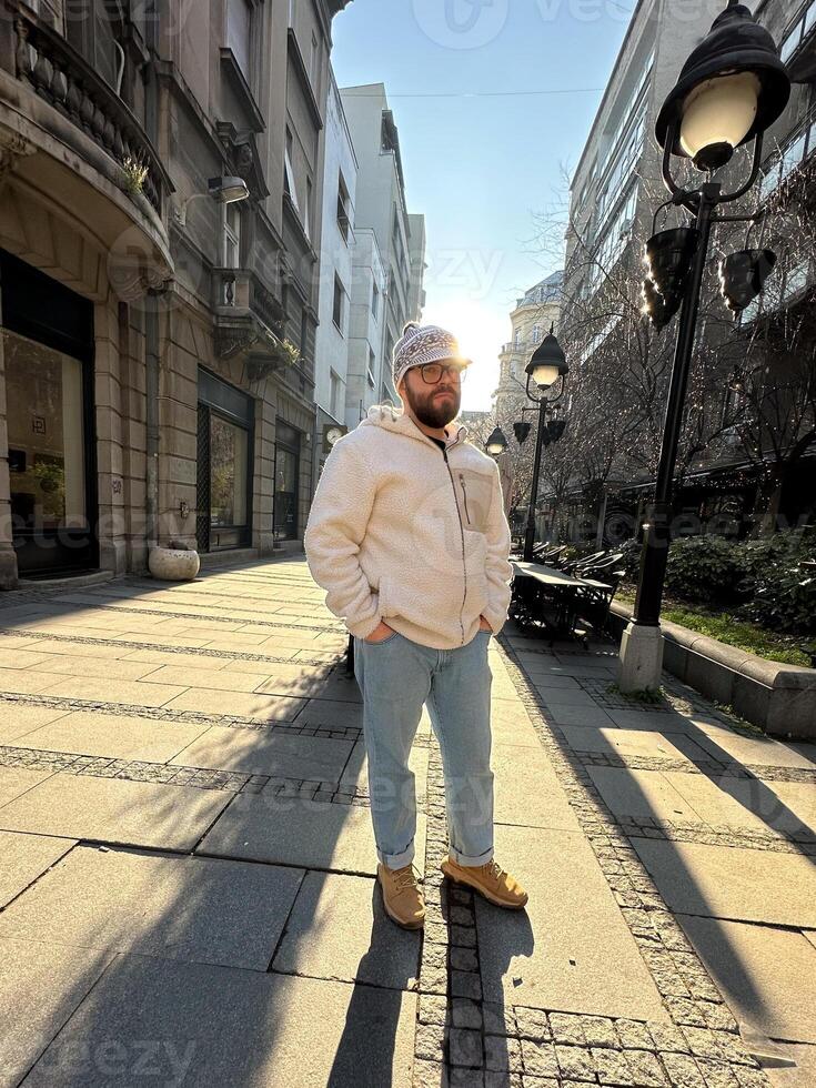 Millennial man stands confidently on a sunlit city street, wearing a cozy jacket and stylish hat. Urban landscape and soft morning light create a scene of casual elegance and modern lifestyle photo