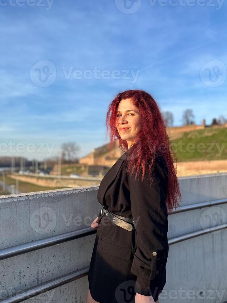 Millennial woman stands confidently in sunlight, with vibrant red hair flowing and dressed in a chic black outfit. Background of a clear blue sky and distant scenery enhances the dynamic photo