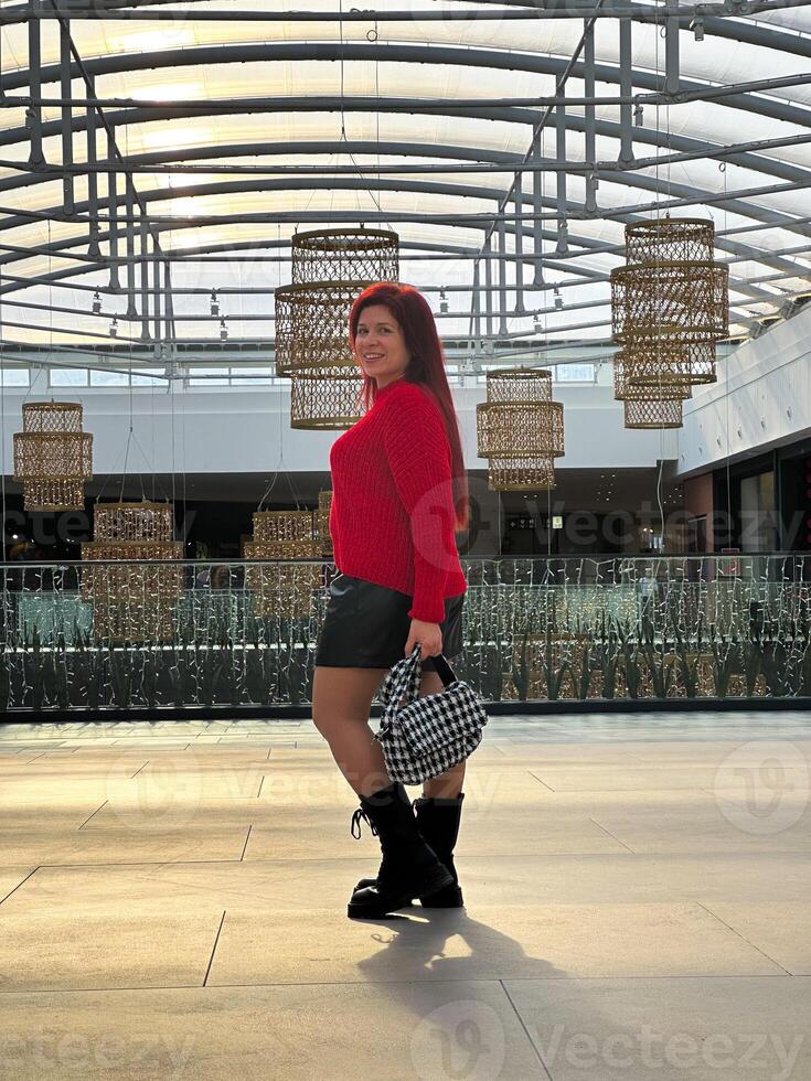 Millennial woman poses confidently in an open, modern environment, wearing a vibrant red sweater and black boots. photo
