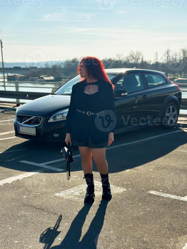 Millennial woman with long curly red hair gazing thoughtfully at the camera on a sunny day. Background features black car and a river, evoking a sense of urban calm. photo