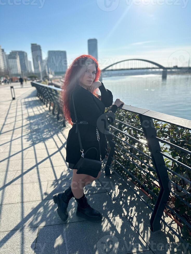 Millennial woman with long curly red hair leans on a black railing, gazing thoughtfully at the camera on a sunny day. Background features modern buildings and a river, evoking a sense of urban calm photo
