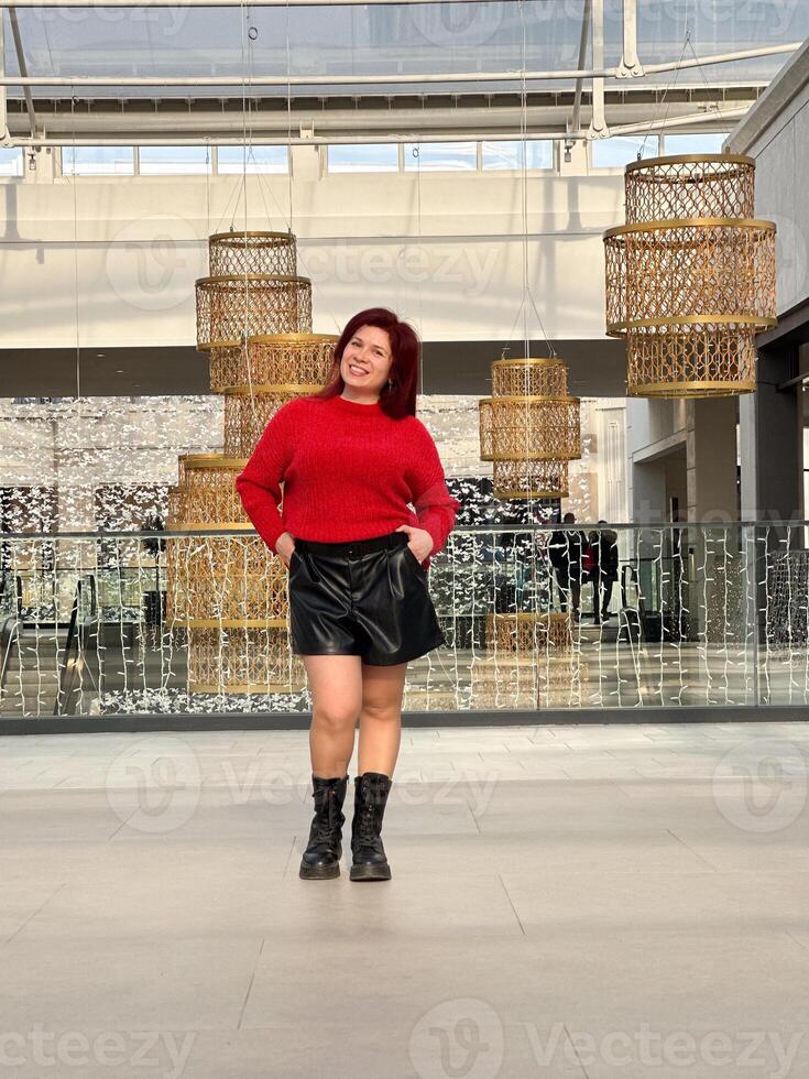 Millennial woman poses confidently in an open, modern environment, wearing a vibrant red sweater and black boots. photo