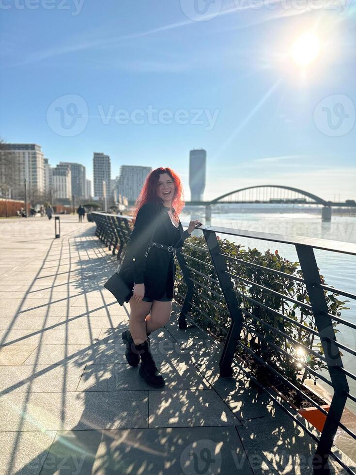 Millennial woman with long curly red hair leans on a black railing, gazing thoughtfully at the camera on a sunny day. Background features modern buildings and a river, evoking a sense of urban calm photo